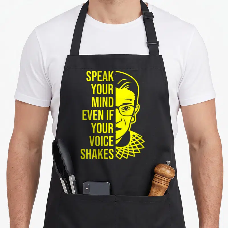 Tablier de cuisine noir avec illustration jaune d'un visage aux lunettes et texteSpeak your mind even if your voice shakes