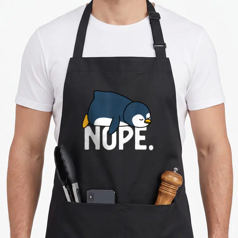 Tablier de cuisine noir avec un pingouin bleu et blanc allongé sur le texte Nope en lettres blanches capitales.