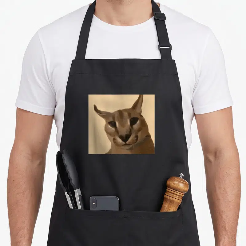 Tablier de cuisine noir avec le portrait pixelisé du chat Big Floppa sur fond beige uni.