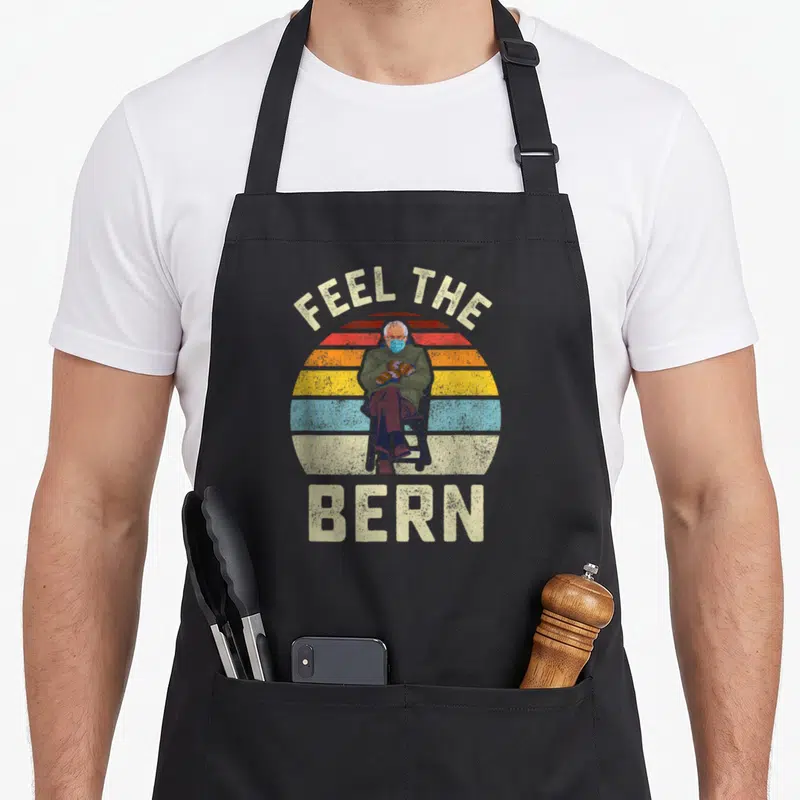 Tablier de cuisine noir avec illustration vintage Feel The Bern et personnage en chaise portant des mitaines.