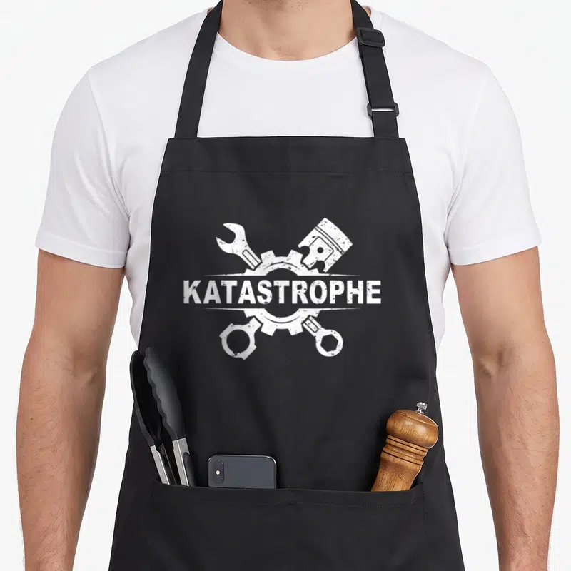 Tablier de cuisine noir avec inscription Katastrophe entourée d'une clé, d'un piston et d'un engrenage en blanc délavé.