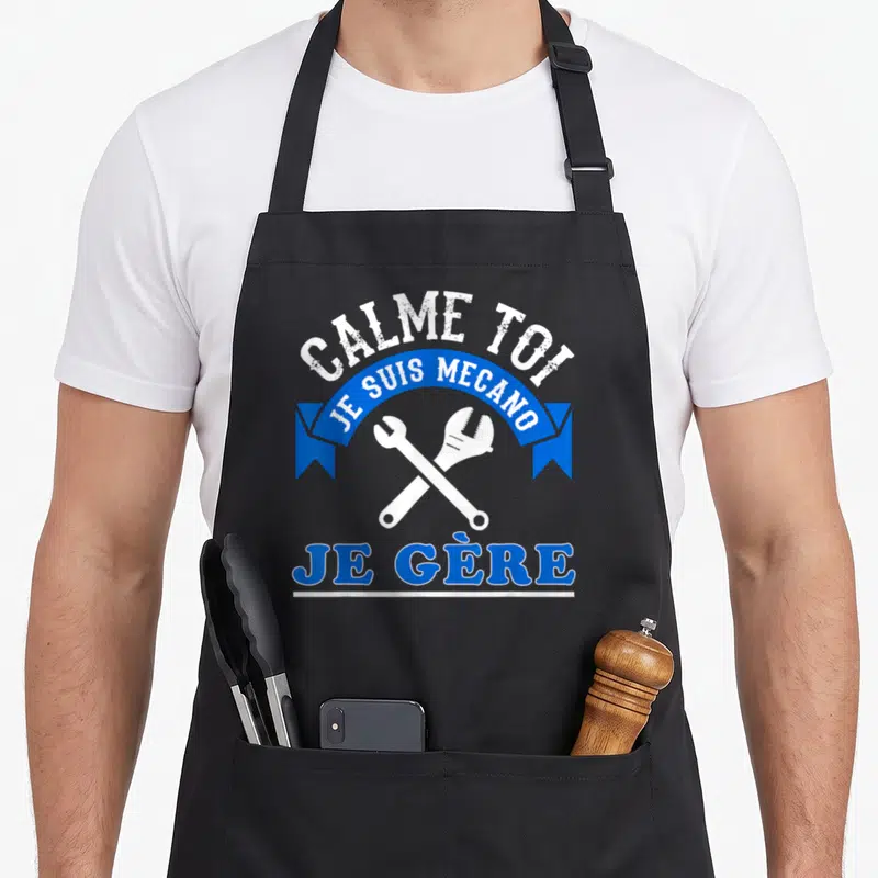 Tablier de cuisine noir avec l'inscription Calme toi je suis mécano je gère et deux clés croisées en blanc sur fond bleu.