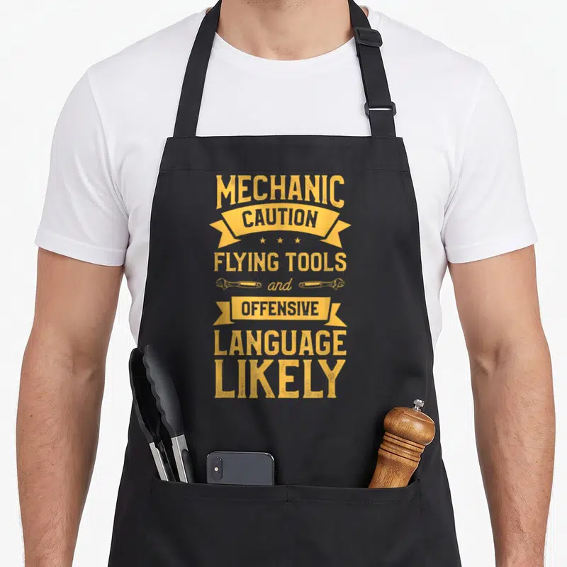 Tablier de cuisine noir avec inscription jaune Caution Flying Tools and Offensive Language Likely et deux clés anglaises.