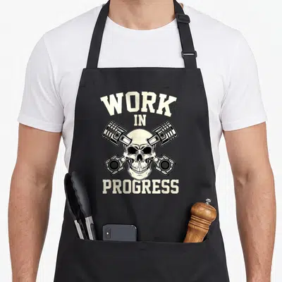 Tablier de cuisine noir avec un crâne et des pistons croisés sur le motif Work in Progress