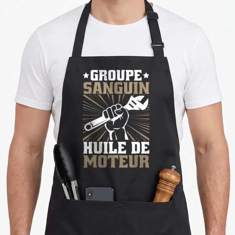 Tablier de cuisine noir avec une main tenant une clé et l'inscription Groupe Sanguin Huile de Moteur en blanc et doré.