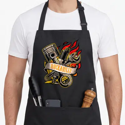 Tablier de cuisine noir avec illustration de piston, clé à molette, flamme et texte ROCKABILLY en bannière.