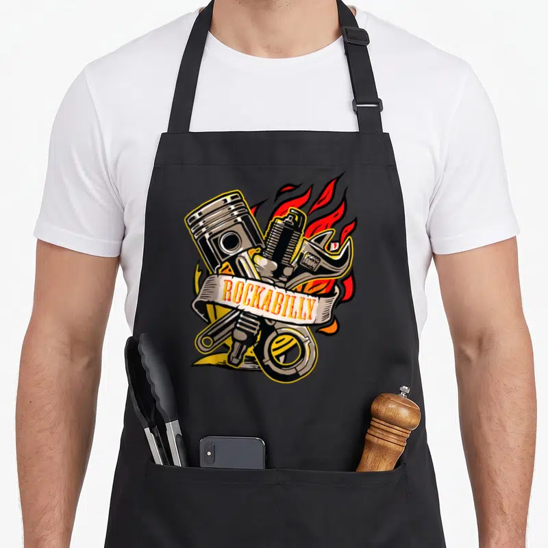 Tablier de cuisine noir avec illustration de piston, clé à molette, flamme et texte ROCKABILLY en bannière.