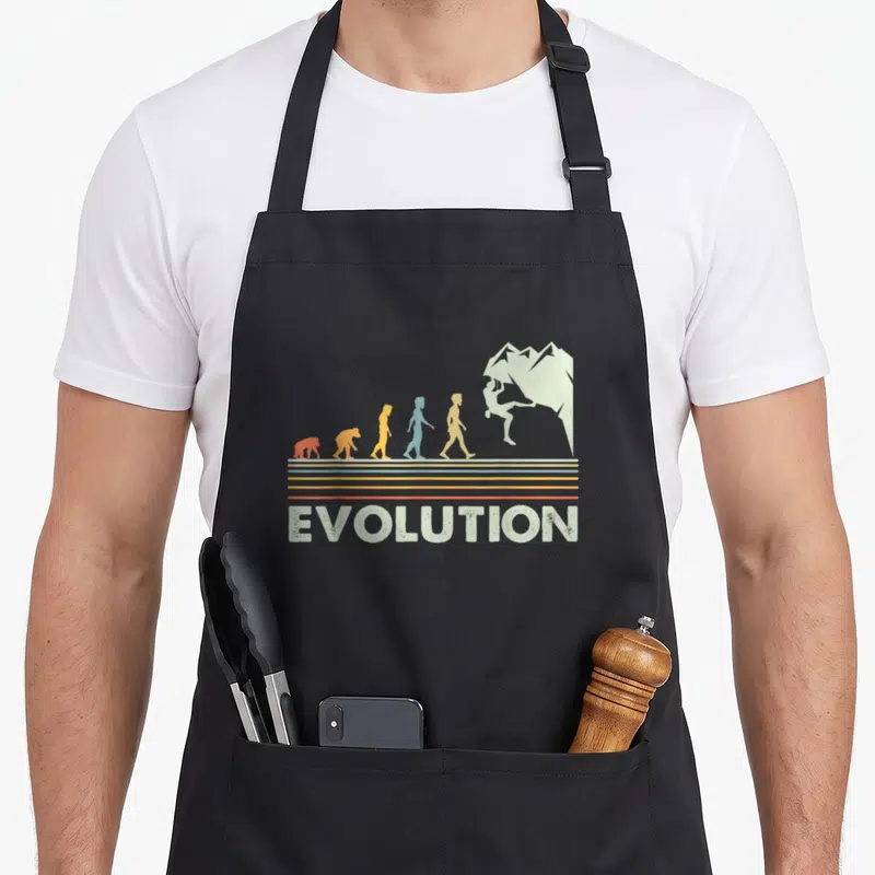 Tablier de cuisine noir avec illustration évolutive montrant des silhouettes colorées menant à un grimpeur sur paroi rocheuse au-dessus du mot Evolution.