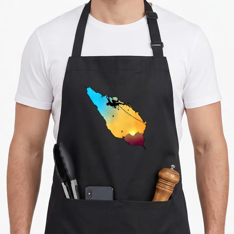 Tablier de cuisine noir avec illustration d'un grimpeur sur une paroi aux couleurs dégradées du soleil couchant.
