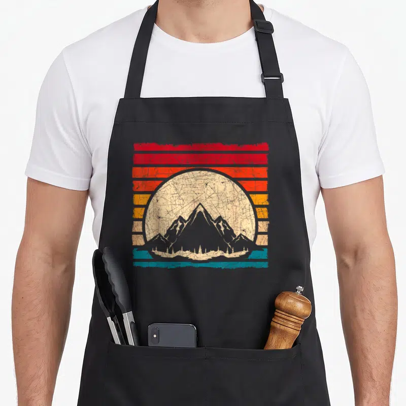 Tablier de cuisine noir avec illustration retro de montagnes et soleil aux couleurs chaudes et rayures horizontales.