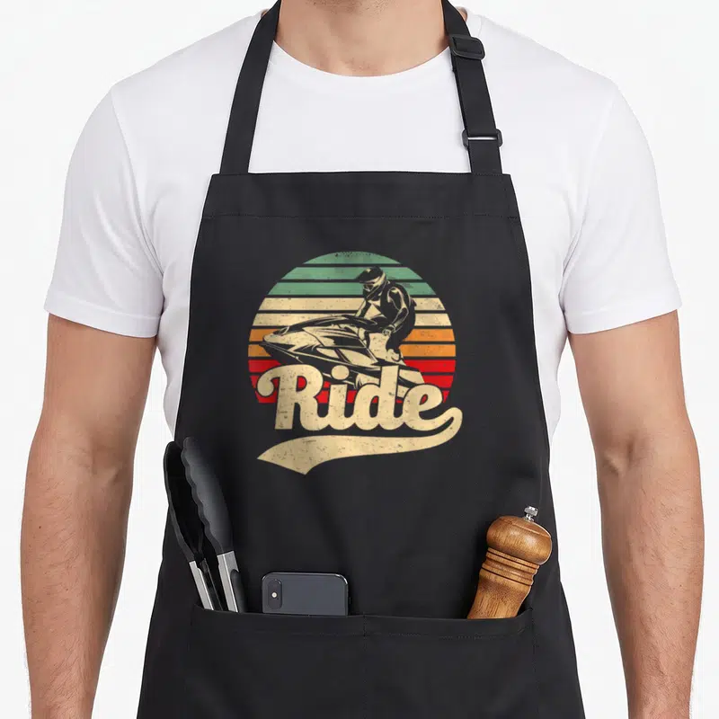 Tablier de cuisine noir avec illustration rétro d'un pilote de jet-ski sur fond de soleil rayé multicolore et le mot Ride en lettres capitales.