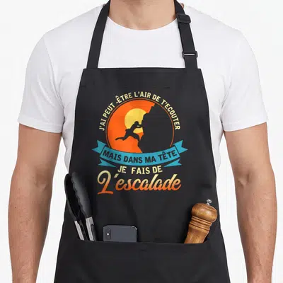 Tablier de cuisine noir avec silhouette d'escaladeur orange et texte humoristique sur fond circulaire bleu et jaune.