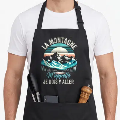 Tablier de cuisine noir avec illustration de montagnes, soleil rayonnant et texte La Montagne M'appelle Je Dois Y Aller en style vintage.