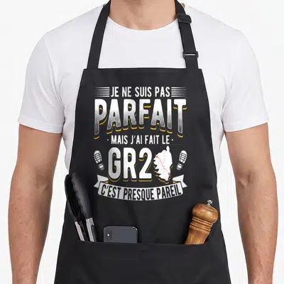 Tablier de cuisine noir avec texte blanc et jaune sur le GR20 et une carte de Corse entre deux traces de chaussures.