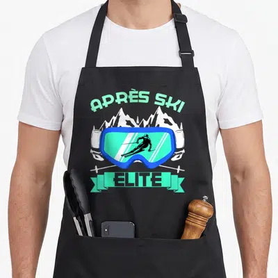 Tablier de cuisine noir avec motif Après Ski Elite, lunettes bleues et silhouettes de montagnes blanches.