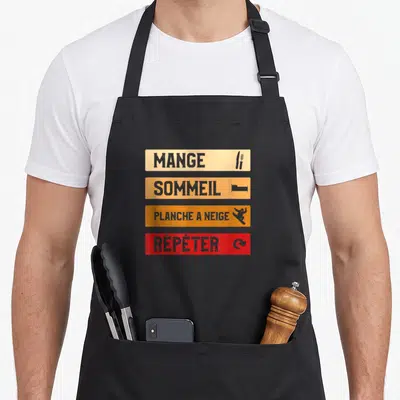 Tablier de cuisine noir avec quatre bandes colorées affichant les mots Mange, Sommeil, Planche à Neige et Répéter accompagnés d'icônes.