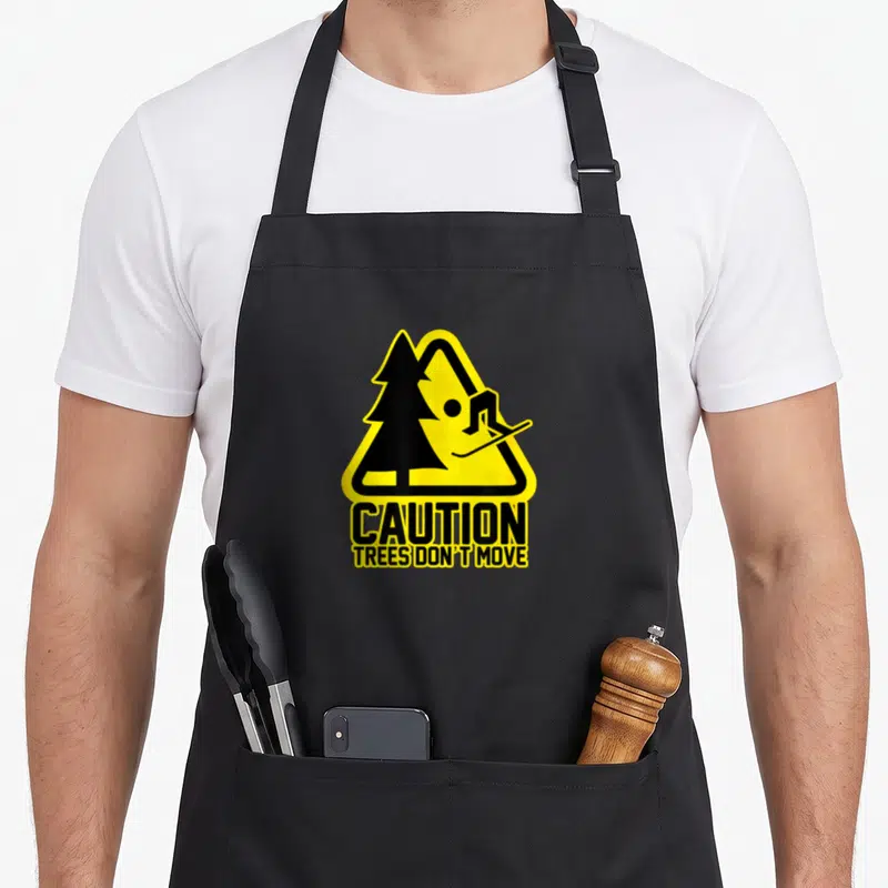 Tablier de cuisine noir avec un panneau jaune signalant un skieur heurtant un arbre, texte Caution Trees Don't Move.