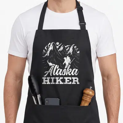 Tablier de cuisine noir avec illustration blanche d'un randonneur et du texte Alaska Hiker devant des montagnes.