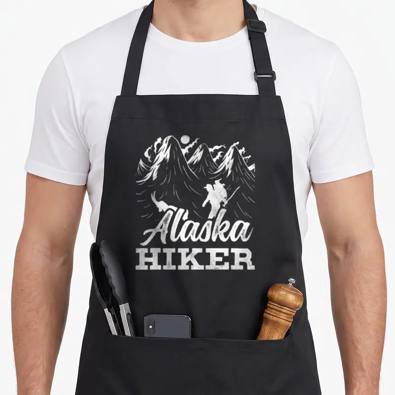 Tablier de cuisine noir avec illustration blanche d'un randonneur et du texte Alaska Hiker devant des montagnes.