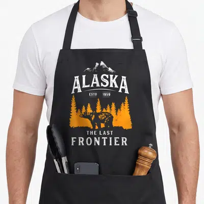 Tablier de cuisine noir avec illustration d'un ours orange, de sapins et du texte Alaska Estp 1959 The Last Frontier en blanc.