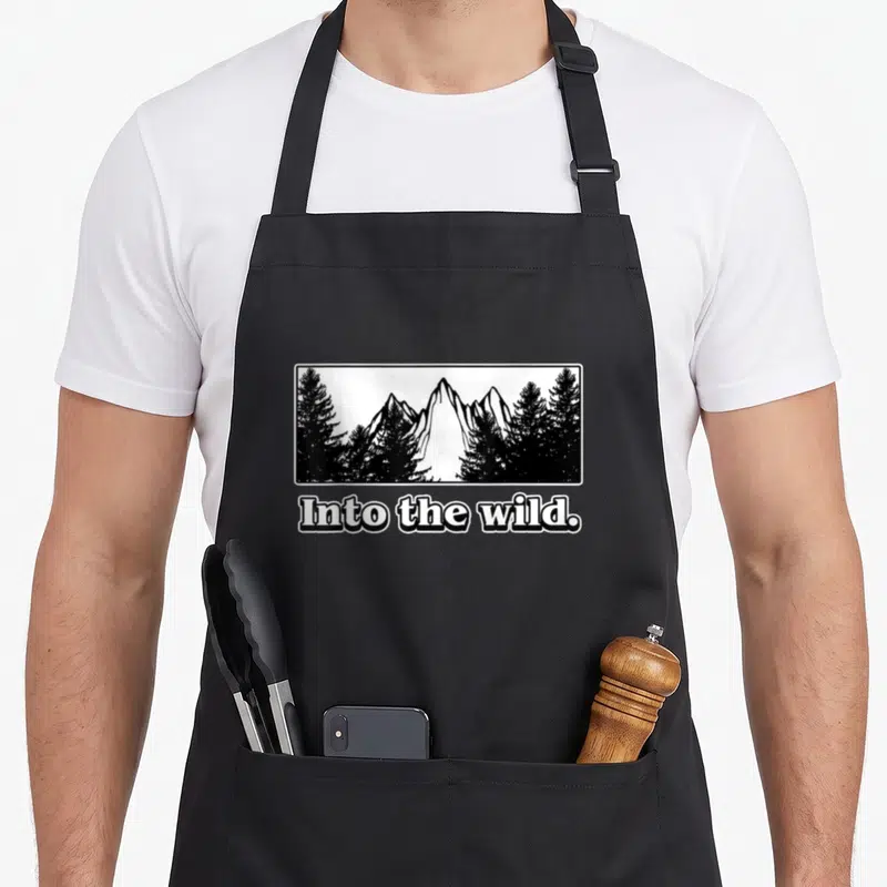 Tablier de cuisine noir avec illustration « Into the wild. » et silhouettes d'arbres et de montagnes en blanc sur fond noir.