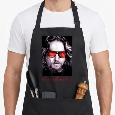Tablier de cuisine noir avec le visage d'un homme aux lunettes rouges et le titre The Big Lebowski en bas.