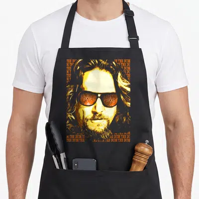 Tablier de cuisine noir avec portrait stylisé jaune et orange de The Dude portant des lunettes de soleil à motifs floraux.