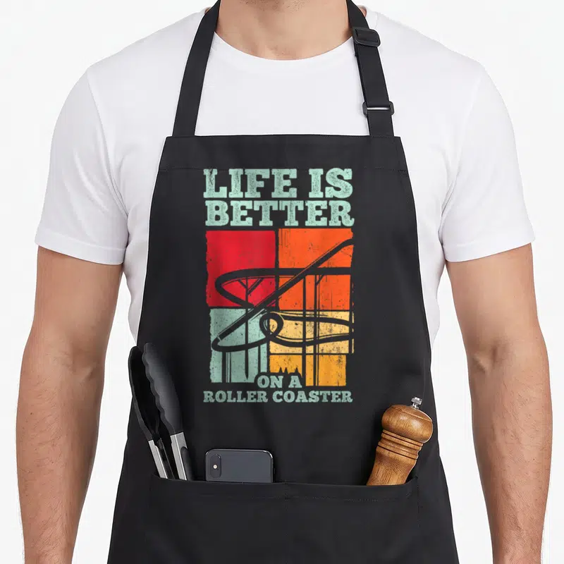 Tablier de cuisine noir avec illustration vintage de montagnes russes et texte Life is Better on a Roller Coaster en pastel et orange.