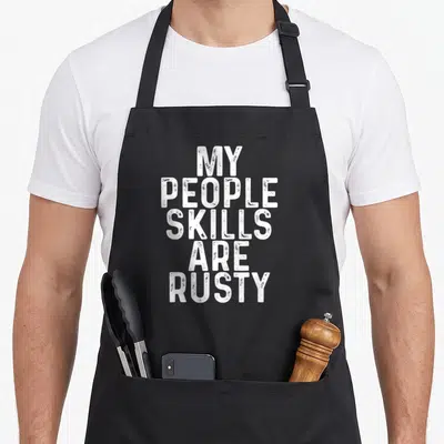 Tablier de cuisine noir avec le texte blanc « Mes compétences sociales sont rouillées » en typographie usée.