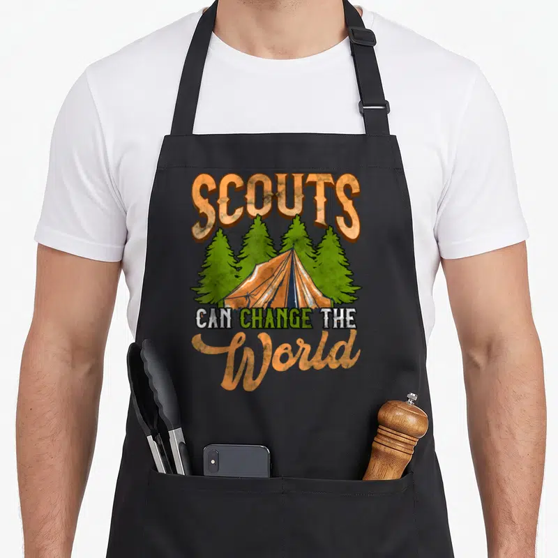 Tablier de cuisine noir avec illustration tente orange, pins verts et texte Scouts can change the World.