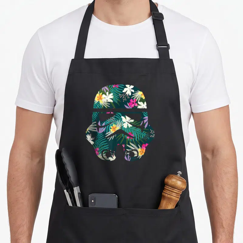 Tablier de cuisine noir avec silhouette de casque de Stormtrooper remplie de fleurs et de feuilles tropicales colorées.