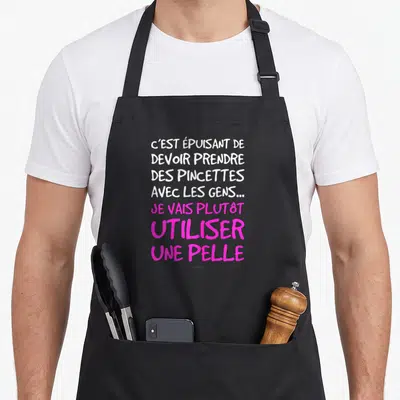 Tablier de cuisine noir avec un texte blanc et rose indiquant qu'il vaut mieux utiliser une pelle que des pincettes avec les gens.