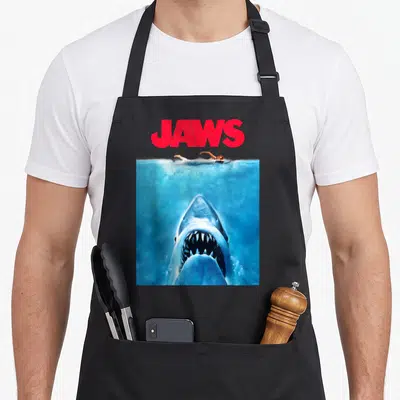 Tablier de cuisine noir avec illustration d'un grand requin sous l'eau et d'une nageuse au-dessus, texte JAWS en rouge.