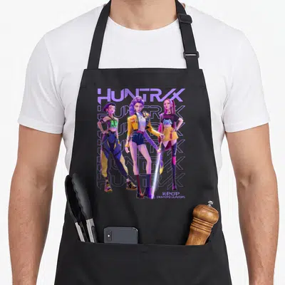 Tablier de cuisine noir avec illustration des trois personnages du groupe Huntrix et le logo stylisé en arrière-plan violet.