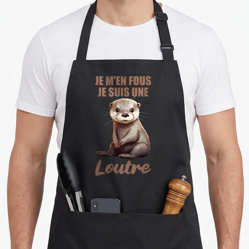 Tablier de cuisine noir avec illustration d'une loutre assise et le texte Je m'en fous je suis une loutre en marron.