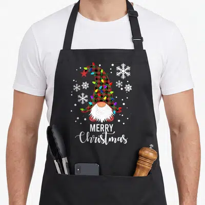 Tablier de cuisine noir avec un gnome à chapeau écossais, guirlandes colorées, flocons de neige et texte Merry Christmas en bas.