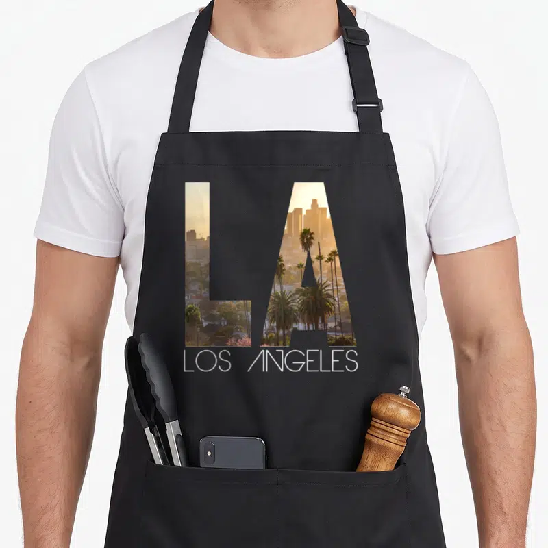 Tablier de cuisine noir avec les lettres LA contenant une photo de palmiers et de ville sous un ciel orangé.