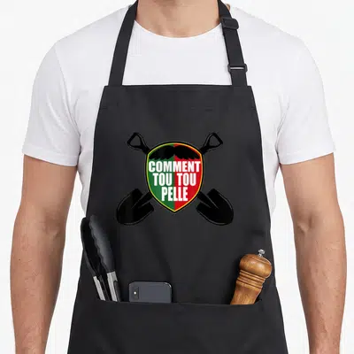 Tablier de cuisine noir avec un écusson vert et rouge affichant « Comment tou tou pelle » et deux pelles noires croisées en arrière-plan.