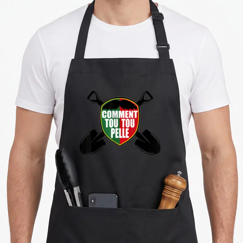 Tablier de cuisine noir avec un écusson vert et rouge affichant « Comment tou tou pelle » et deux pelles noires croisées en arrière-plan.