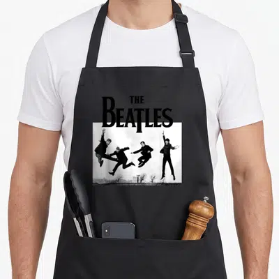 Tablier de cuisine noir avec l'inscription The Beatles et quatre hommes en train de sauter en air sur un fond blanc.