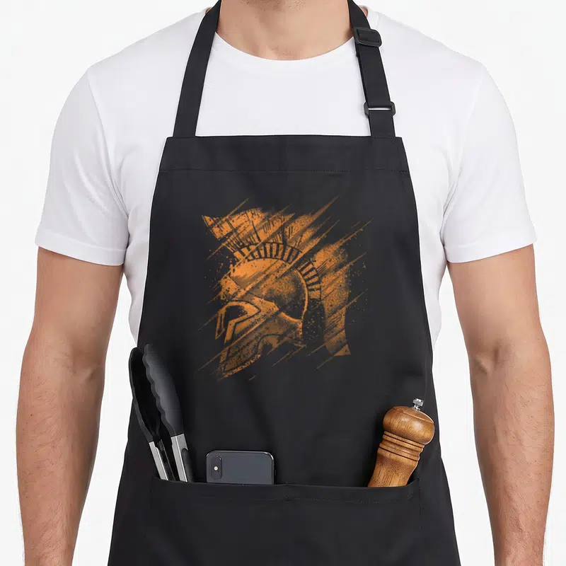 Tablier de cuisine noir avec illustration orange d'un casque de guerrier spartan aux traits dynamiques et texture pointilliste.