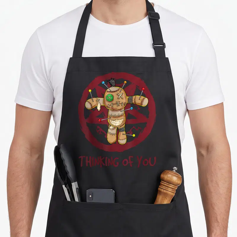 Tablier de cuisine noir avec une poupée vaudou aux épingles colorées et l'inscription THINKING OF YOU en bas.