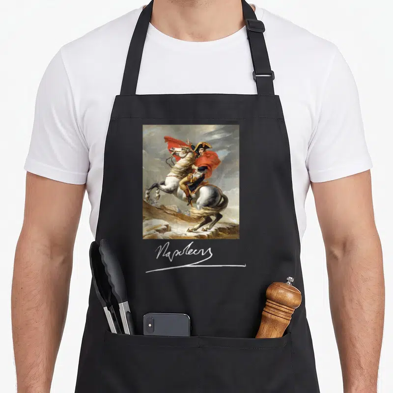 Tablier de cuisine noir avec reproduction de Napoléon Bonaparte chevauchant un destrier blanc sous un ciel nuageux, signature blanche en dessous.