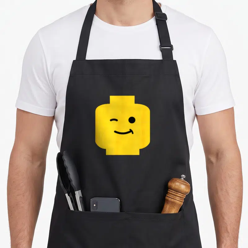 Tablier de cuisine noir avec un grand visage jaune minifigurine clignant de l'œil et souriant