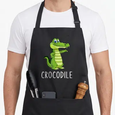 Tablier de cuisine noir avec un crocodile vert mignon debout et le mot crocodile écrit en blanc en dessous.