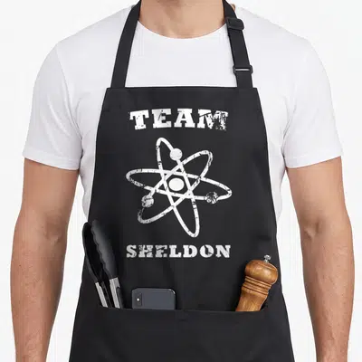 Tablier de cuisine noir avec le motif blanc Team Sheldon et un symbole atomique stylisé au centre.