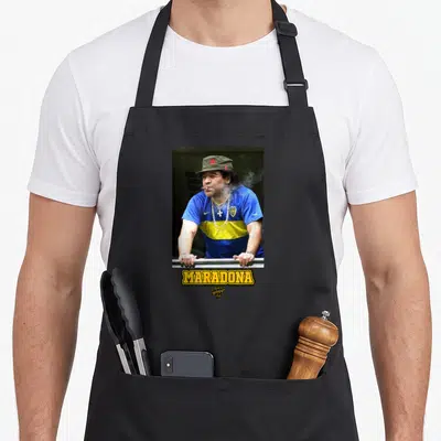 Tablier de cuisine noir avec Maradona en maillot bleu et jaune fumant un cigare sous une casquette verte.