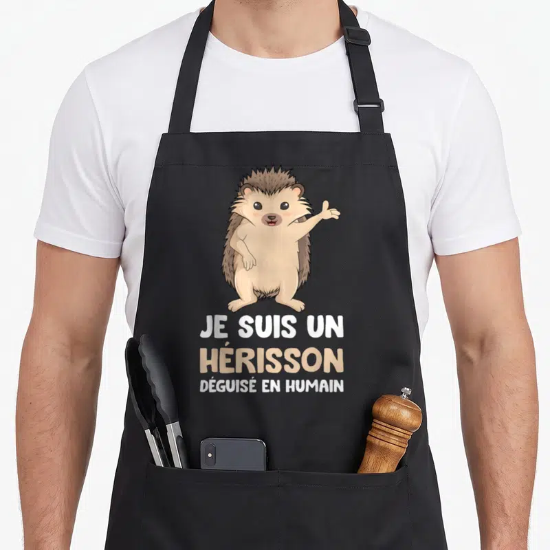 Tablier de cuisine noir avec un hérisson marron debout et le texte Je suis un hérisson déguisé en humain imprimé en blanc et beige.