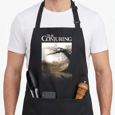Tablier de cuisine noir avec une maison hantée et un arbre sec portant une corde.