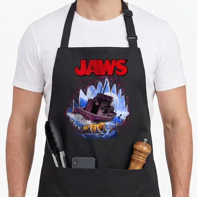 Tablier de cuisine noir avec illustration Jaws montrant un bateau entre les dents d'une orque dans des eaux bleues.